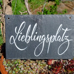 Op de afbeelding: Een zwart leisteen bord met witte letters die "Lieblingsplatz" lezen. Het bord hangt aan een houten tak en is omringd door groene planten.