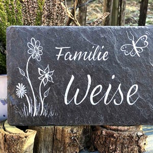 Op de afbeelding: Een rechthoekige donkergrijze leistenen plaat met witte gegraveerde tekst "Familie Weise". Het ontwerp bevat een witte illustratie van drie bloemen aan de linkerkant en een vlinder in de rechterbovenhoek van het stenen oppervlak.