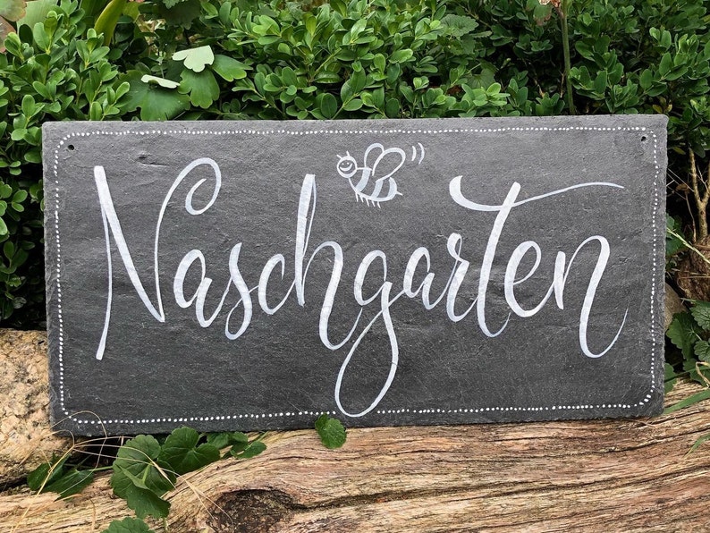 Op de afbeelding: Een zwart leisteenbord met witte letters met de tekst "Naschgarten" met een kleine bij-illustratie. Het bord heeft een witte gestippelde rand.