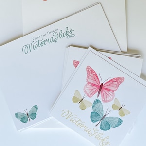 Może przedstawiać: Biały zestaw artykułów papierniczych z ilustracjami motyli w kolorze różowym, niebieskim i żółtym. Zestaw zawiera notatniki i karty z imieniem "Victoria Glicks" w eleganckim skrypcie. Jeden notatnik głosi "From the desk of Victoria Glicks".