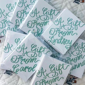 以下が含まれることがあります： 白地のギフトカードのコレクション。ターコイズ色の筆記体で「A Gift From」と書かれています。カードは個別に透明なプラスチックで包装されています。「Carol」や「Lindsey」などの名前が書かれています。