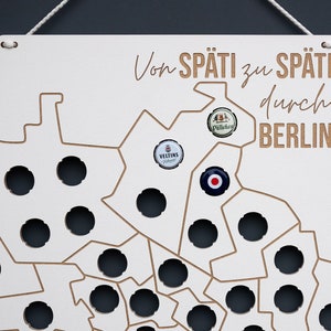 Könnte beinhalten: Eine weiße Holzkarte von Berlin mit dem Text "Von Späti zu Späti durch Berlin" in brauner Schrift. Die Karte hat mehrere Löcher für Flaschenverschlüsse, und drei Flaschenverschlüsse sind auf der Karte angebracht.