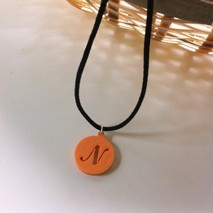 May include: Orange pendant necklace with a black cord. The pendant is a circular shape with the letter "N" cut out. The necklace is hanging in front of a white surface and a wicker basket.
