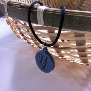 May include: A dark gray pendant necklace with a black cord. The pendant is circular with a stylized letter "Y" cut out of the center. The necklace is displayed near a woven basket.