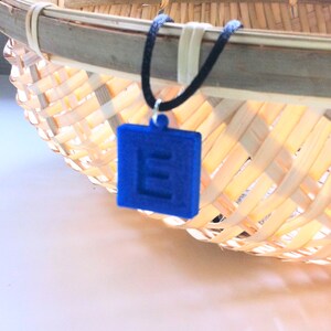 May include: A blue square pendant necklace with the letter "E" cut out. The pendant hangs from a black cord. The background features a woven basket.