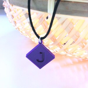 May include: A purple pendant necklace with a black cord. The pendant is a diamond shape with the letter "J" in the center. The necklace is hanging in front of a woven basket.