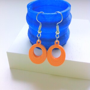 May include: A pair of orange hoop earrings with silver-colored hooks are displayed in front of a blue, textured cylindrical object. The earrings are circular with a hollow center. The blue object has a layered design and sits on a white surface.