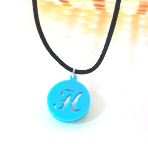 May include: A bright turquoise pendant necklace featuring a cut-out letter "K" within a circular frame. The pendant hangs from a black cord necklace. The necklace is displayed against a white background.