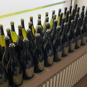 Puede incluir: Una gran colección de botellas de champán de vidrio verde oscuro dispuestas en filas. Cada botella presenta una etiqueta de color crema con texto negro y un emblema en forma de escudo. Las botellas tienen cuellos largos y delgados.