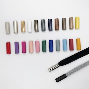 Puede incluir: Una colección de veintidós herretes cilíndricos de metal en una variedad de colores sólidos, incluyendo metálicos, blanco, negro, rojo, azul, verde, rosa y amarillo, junto a dos cordones, uno negro y otro gris, con herretes de metal acoplados.