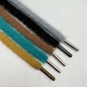 May include: Close-up of five fuzzy shoelaces in various colors: mustard yellow, teal, brown, black, and gray. Each lace has a metal aglet at the end. The laces are arranged diagonally against a white background.