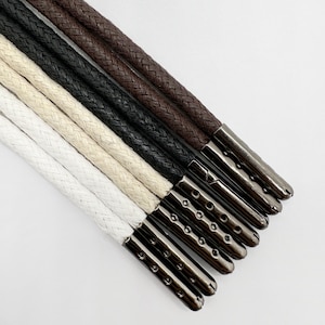 May include: A set of six braided cords in white, beige, black, and dark brown. Each cord is finished with a metallic gunmetal-colored aglet featuring a series of small circular indentations along the side, providing a secure and decorative end.