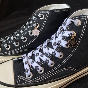 May include: A pair of black high-top canvas trainers with white laces and a white floral print. The laces are tied with a decorative charm featuring a white flower.