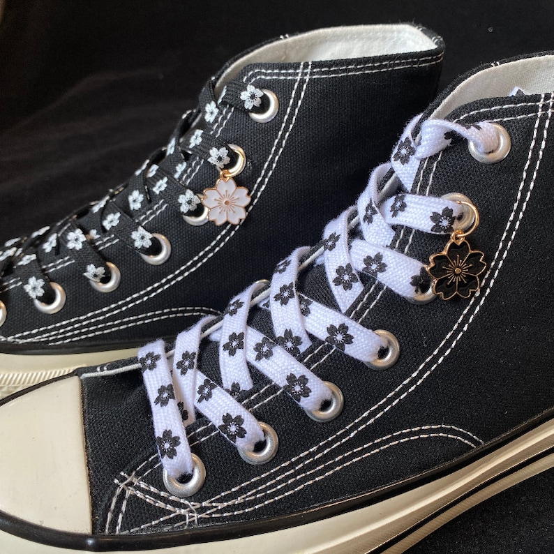 May include: A pair of black high-top canvas trainers with white laces and a white floral print. The laces are tied with a decorative charm featuring a white flower.
