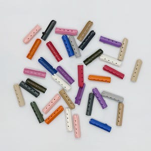 Könnte beinhalten: Eine Sammlung bunter, zylindrischer Haarwickler in verschiedenen Farbtönen, darunter Rosa, Orange, Grün, Blau und Lila. Jeder Wickler hat eine Reihe kleiner Löcher und ein gebogenes Design. Die Wickler sind auf einer weißen Oberfläche verstreut.