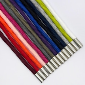 Puede incluir: Una colección de doce cordones trenzados en varios colores, incluyendo blanco, verde lima, gris, negro, azul, morado, gris oscuro, rosa, rojo, naranja, rosa claro y rojo oscuro, cada uno terminado con una punta de metal plateado pulido.