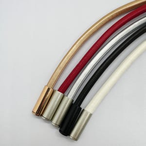 Pode incluir: Cinco cordões finos e arredondados em dourado, vermelho, prateado, preto e branco, cada um com uma superfície texturizada. Cada cordão é finalizado com uma ponteira metálica cilíndrica e lisa em tons metálicos a condizer ou contrastantes.