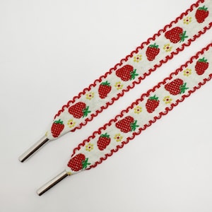 Könnte beinhalten: Zwei Schnürsenkel vor weißem Hintergrund. Sie weisen ein sich wiederholendes Muster aus roten Erdbeeren und gelben Blumen auf. Die Ränder sind mit einem roten, gewellten Rand versehen. Die Aglets sind silberfarben.