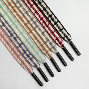 Puede incluir: Una colección de cordones de zapatos coloridos con un patrón de pata de gallo. Los cordones son de varios colores, incluyendo rosa, morado, azul, marrón, rojo y negro. Cada cordón tiene una puntera de plástico negro.