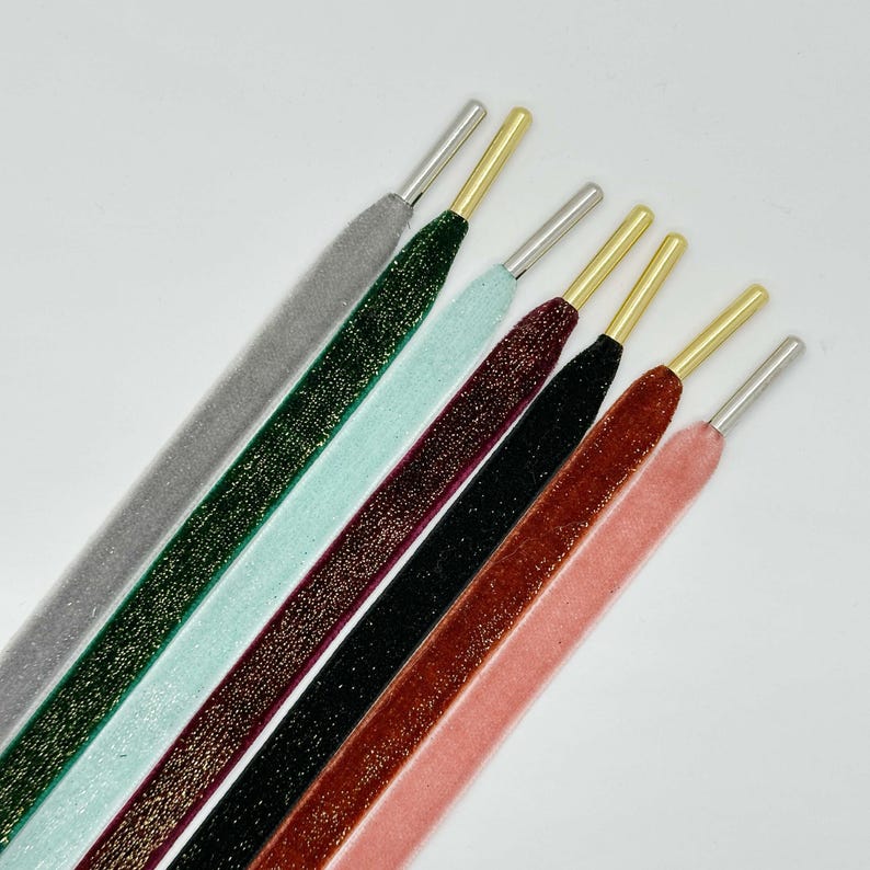 May include: A set of six shoelaces in various colors, including gray, green, mint, burgundy, black, and orange. Each shoelace has a gold-colored metal tip and a silver-colored metal aglet.