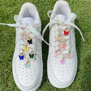 Könnte beinhalten: Weiße Sneaker mit schillernden Schnürsenkeln und Schmetterlings-Schuhschmuck in verschiedenen Farben, darunter Blau, Grün, Gelb, Rosa und Rot.