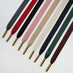 Könnte beinhalten: Eine Auswahl an farbigen Schnürsenkeln mit goldfarbenen Ösen. Die Schnürsenkel sind in verschiedenen Farben erhältlich, darunter Dunkelgrau, Rot, Schwarz, Rosa, Beige, Weiß, Marineblau, Grün und Braun. Jede Schnürsenkel hat ein strukturiertes Muster.