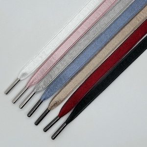Puede incluir: Una colección de cordones planos en varios colores, incluyendo blanco, rosa, gris, azul, beige, rojo y negro. Cada cordón tiene una puntera metálica plateada o negra. Los cordones están dispuestos diagonalmente, mostrando sus diferentes tonos y texturas.