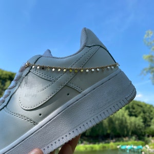 Puede incluir: Zapatillas blancas de cuero con una cadena de estrellas doradas alrededor del tobillo.