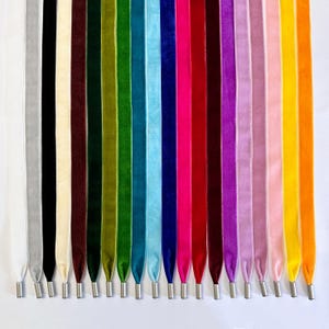 May include: A collection of colorful velvet ribbons in various shades, including silver, black, cream, burgundy, green, blue, pink, red, purple, and yellow. Each ribbon has a metal aglet at the end. The ribbons are arranged vertically against a white background.