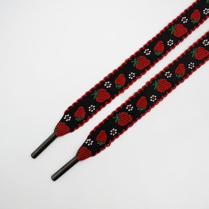 May include: Two black shoelaces with a repeating pattern of red strawberries and white flowers. The edges are scalloped with a red border. The aglets are dark gray.