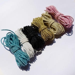Puede incluir: Seis paquetes de cordón metálico dispuestos en una fila diagonal. Los colores incluyen azul cerceta, blanco, negro, dorado, oro claro y rosa. Cada paquete consiste en una cuerda delgada, brillante y trenzada en un bucle ovalado compacto.