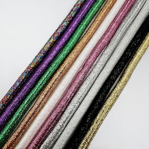 Puede incluir: Una colección de cordones brillantes y coloridos en varios tonos, incluyendo arcoíris, morado, verde, dorado, blanco, rosa, plateado, negro y dorado. Los cordones están dispuestos en diagonal, mostrando sus superficies texturizadas.