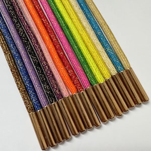 Puede incluir: Una colección de cordones de zapatos coloridos con terminales metálicos dorados. Los cordones son de varios colores, incluyendo dorado, azul, morado, negro, rojo, naranja, rosa y verde. Los cordones están tejidos apretados y tienen un ligero brillo.