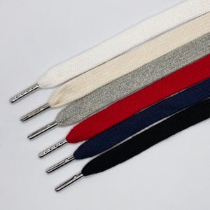 Puede incluir: Un juego de seis cordones planos para calzado en blanco, crema, gris, rojo, azul marino y negro. Cada cordón presenta un herrete metálico plateado y liso en el extremo, aportando un acabado limpio a la textura tejida de los cordones alineados.