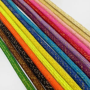 Könnte beinhalten: Eine Ansammlung von parallel diagonal angeordneten Kordeln, die jeweils eine geflochtene Textur mit eingewebten metallischen Goldfäden aufweisen. Die Kordeln zeigen ein lebendiges Farbspektrum, darunter Grün, Braun, Orange, Gelb, Blau, Violett, Rot, Pink und Gold.