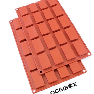 May include: Two orange silicone baking moulds with 24 rectangular cavities. The moulds are stacked on top of each other. The bottom mould has the word "OGGIBOX" printed on it.