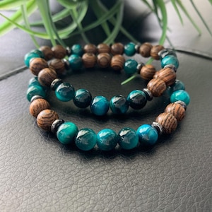 May include: Two beaded bracelets. One bracelet features teal and black beads, and the other has wooden beads. Both bracelets have a mix of dark metal beads. The bracelets are displayed on a black surface.
