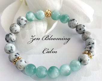 May include: A beaded bracelet featuring alternating light blue and speckled gray beads, with gold-colored accents. The bracelet has the text "Zen Blooming Calm" and "Find Stillness in Every Moment".