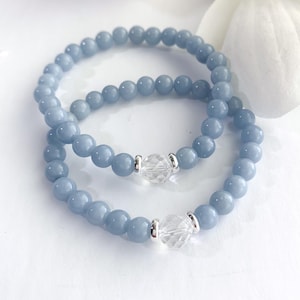 May include: Two light blue beaded bracelets with a clear, faceted crystal accent. The bracelets are made of small, round beads and are displayed on a white surface. A white flower is in the background.