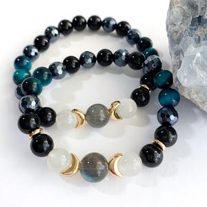 May include: Two beaded bracelets. One bracelet features black, teal, and gray beads. The other bracelet has white, gray, and black beads with gold-colored crescent moon accents. The bracelets are on a white background.
