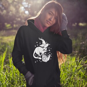 May include: Black hooded sweatshirt featuring a white graphic of a cat wearing a witch's hat and holding a book with stars and planets around it.