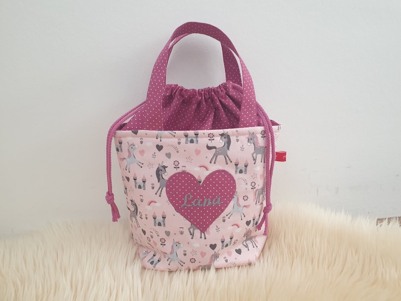 Puede incluir: Bolsa de cord&oacute;n rosa con un coraz&oacute;n rosa que dice "Lana". La bolsa tiene un estampado de unicornios rosa y blanco con castillos y corazones.