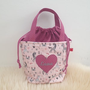 Puede incluir: Bolsa de cord&oacute;n rosa con un parche en forma de coraz&oacute;n rosa que dice "Lana" y un estampado de unicornio. La bolsa tiene un forro de lunares rosas y un asa rosa.
