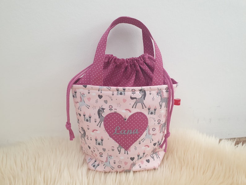 Puede incluir: Bolsa de cord&oacute;n rosa con un parche en forma de coraz&oacute;n rosa que dice "Lana" y un estampado de unicornio. La bolsa tiene un forro de lunares rosas y un asa rosa.