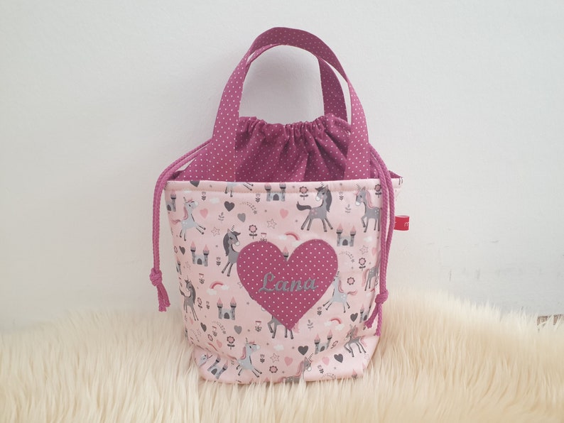 Puede incluir: Bolsa de cord&oacute;n rosa con estampado de unicornio y castillo. La bolsa tiene un coraz&oacute;n rosa con el nombre "Lana" bordado.