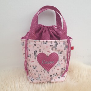 Puede incluir: Bolsa de cord&oacute;n rosa con un parche en forma de coraz&oacute;n rosa que dice "Lana" y un estampado de unicornio. La bolsa tiene asas de lunares rosas.