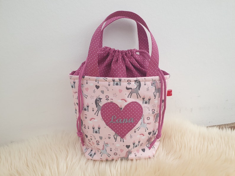 Puede incluir: Bolsa de cord&oacute;n rosa con un parche en forma de coraz&oacute;n rosa que dice "Lana" y un estampado de unicornio. La bolsa tiene asas de lunares rosas.