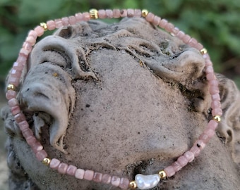 Könnte beinhalten: Ein zartes Armband aus rosa Edelsteinen mit goldenen Akzenten und einer einzelnen weißen Perle. Das Armband besteht aus kleinen, quadratisch geschliffenen rosa Steinen und ist mit kleinen goldenen Perlen durchsetzt. Das Armband ist etwa 18 cm lang.