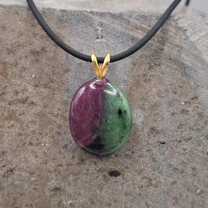 Könnte beinhalten: Ein Anhänger mit einem polierten, ovalen Zoisit-Stein, der deutliche Abschnitte in tiefem Magenta und Waldgrün aufweist. Der Stein wird von einer goldfarbenen Metallöse gehalten und hängt an einer schlichten, glatten schwarzen Kordelkette.
