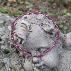 Könnte beinhalten: Ein zartes Perlenarmband mit kleinen, facettierten, dunkelrosa Edelsteinperlen in kreisförmiger Anordnung, durchsetzt mit winzigen, glatten, goldfarbenen Metall-Abstandshaltern für ein subtiles und raffiniertes Erscheinungsbild.